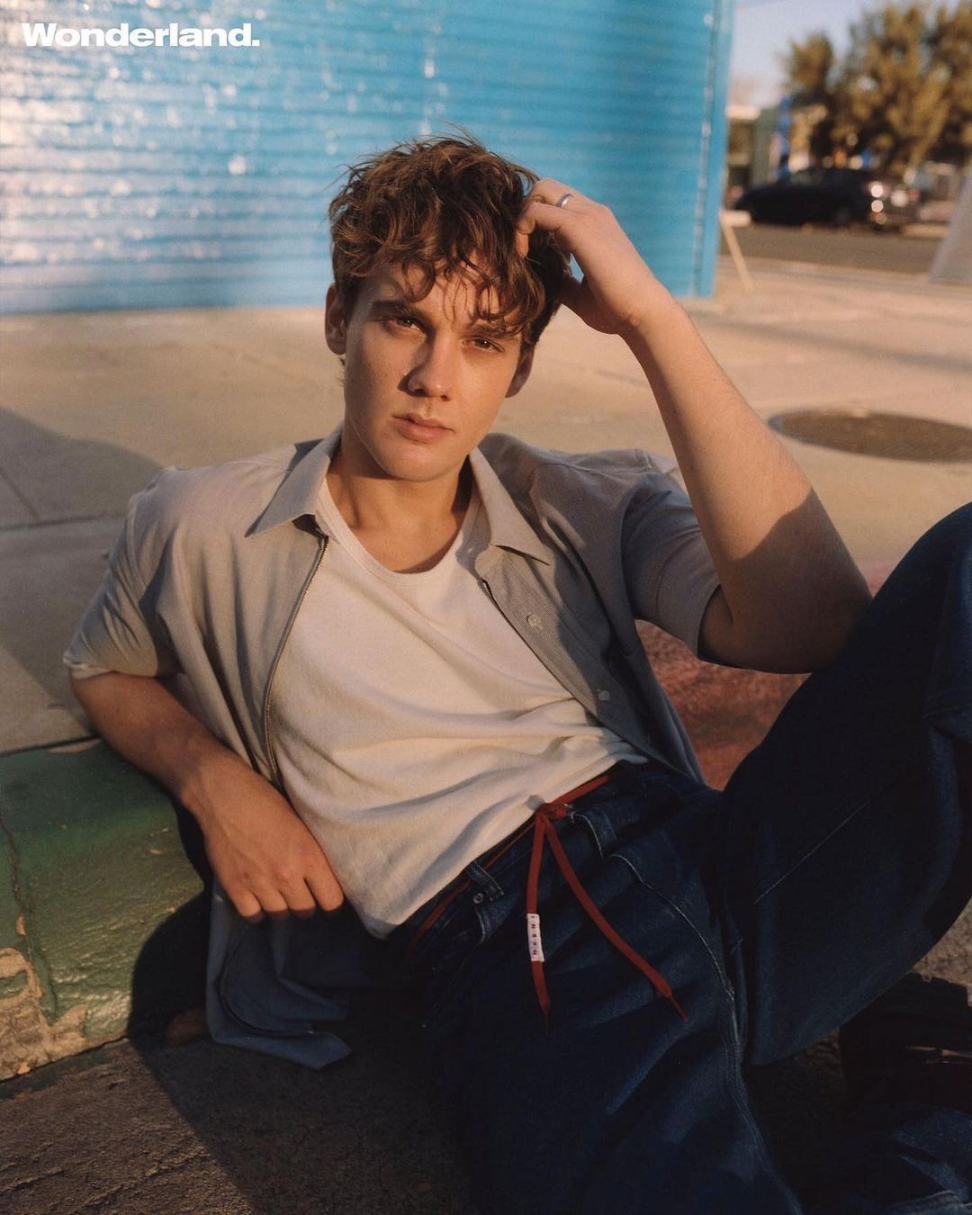 Man with curly brown hair in an unbuttoned grey shirt over a white tee, posing outdoors in blue jeans against a textured blue background.