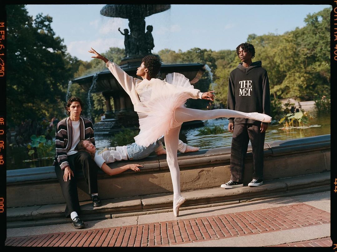 Outdoor shot of four young adults in a park; a ballerina in a pink tutu stands en pointe while others pose against a stone wall near a fountain.