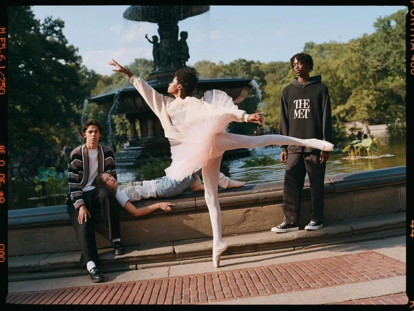Outdoor shot of four young adults in a park; a ballerina in a pink tutu stands en pointe while others pose against a stone wall near a fountain.
