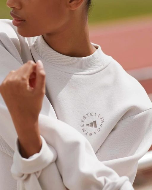 Woman wearing a light grey Stella McCartney Adidas sweatshirt with the logo visible, outdoors on a track in a medium shot.