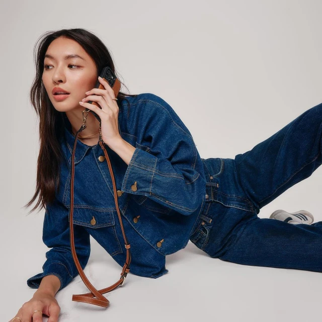 Young woman in a denim jacket and jeans lies on a white background, holding a phone with a brown case attached to a brown strap.