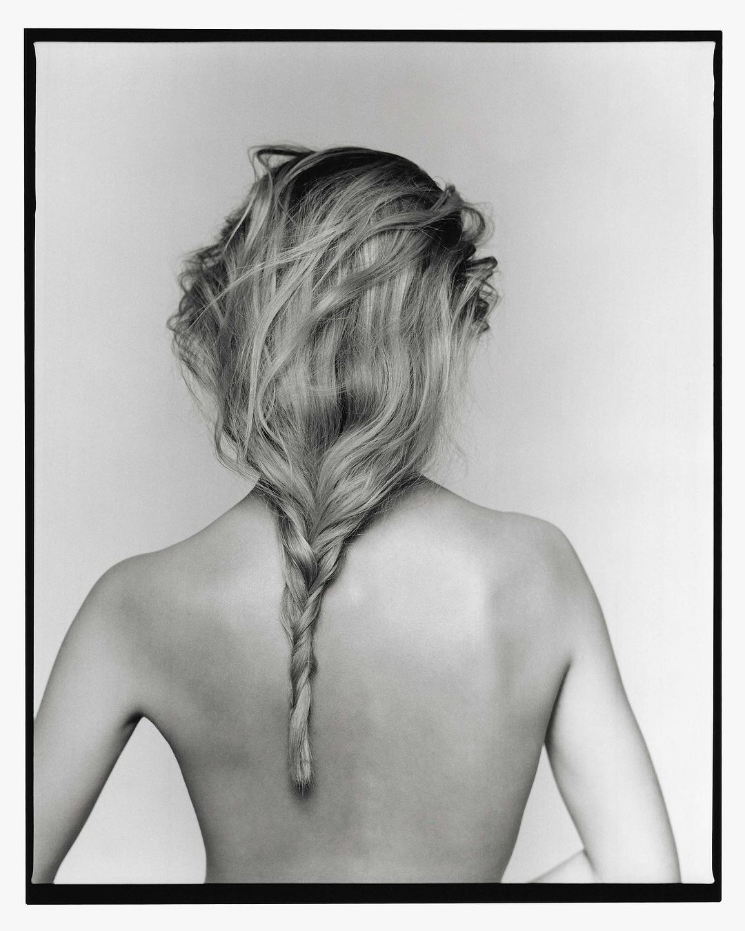 Back view of a blond woman with a braided hairstyle, against a plain light gray background in a black-and-white studio photograph.