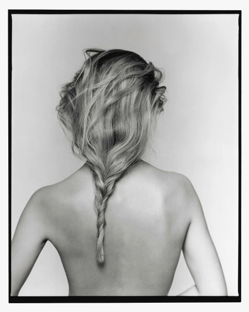 Back view of a blond woman with a braided hairstyle, against a plain light gray background in a black-and-white studio photograph.