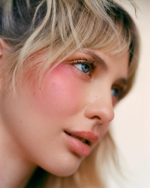 Close-up portrait of a blonde woman with blue eyes wearing pink blush on her cheeks and pink lipstick, with soft lighting and a light backdrop.