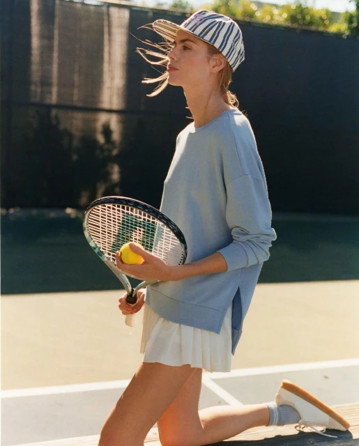 Adult female on tennis court kneeling and holding racquet with tennis ball; wearing sweatshirt, skirt, baseball cap, socks and sneakers.
