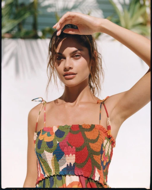 Attractive woman in a floral patterned dress poses outdoors with sunlight casting palm tree shadows on a white wall behind her.