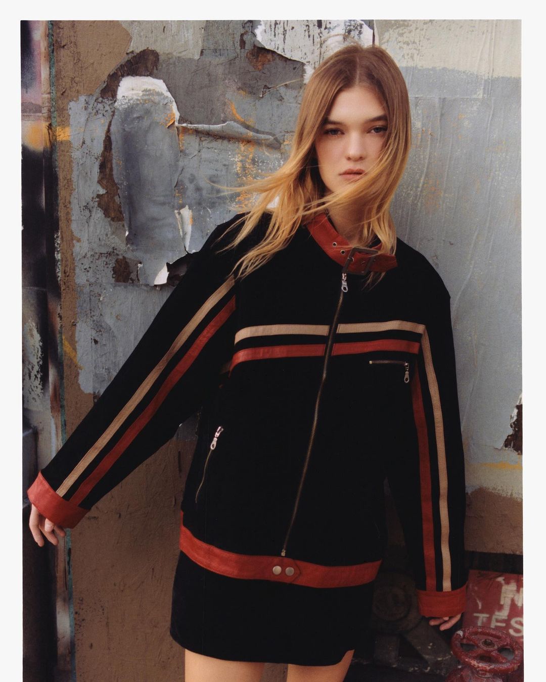 Young woman with long brown hair in a black jacket with brown and red stripes posing against a textured urban wall on a cloudy day.