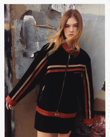 Portrait of a female model wearing a black jacket with red and beige stripes standing against a textured urban wall