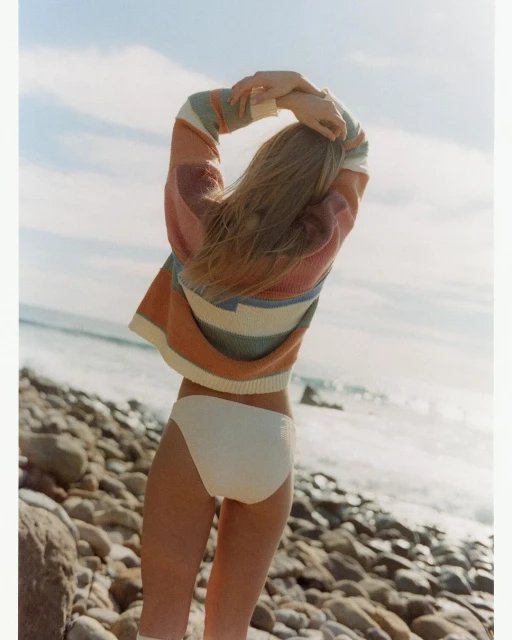 A woman stands on a rocky beach with a pastel striped sweater and white bikini bottom, gazing out at the ocean under a blue sky.