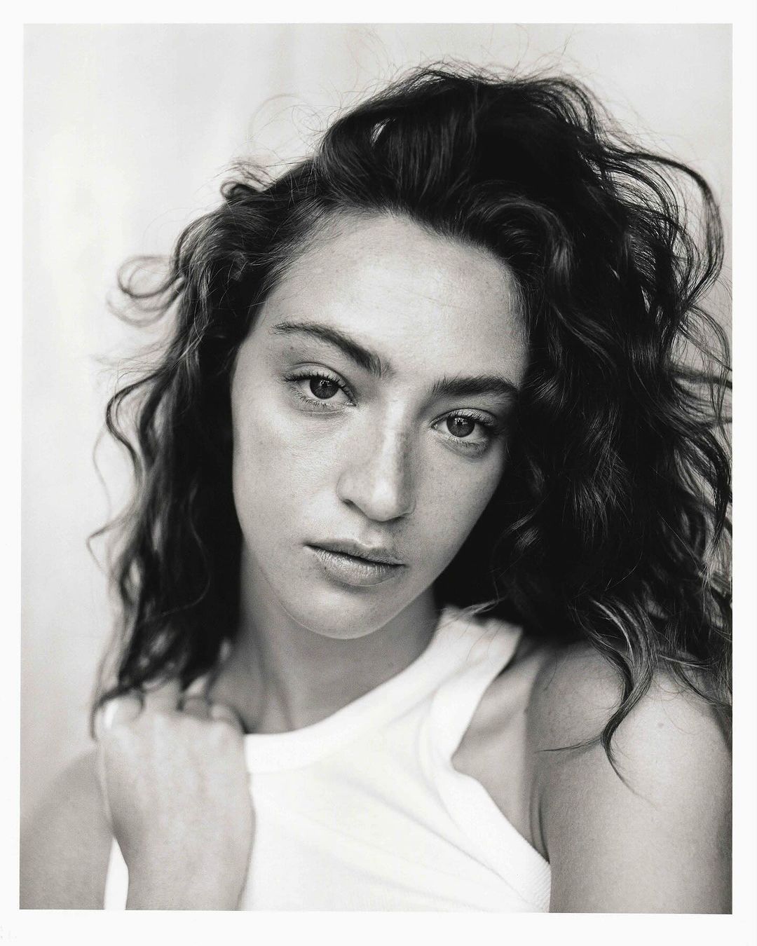 Monochrome portrait of a young adult woman with curly hair and a white top, looking at the camera against a blurred background.