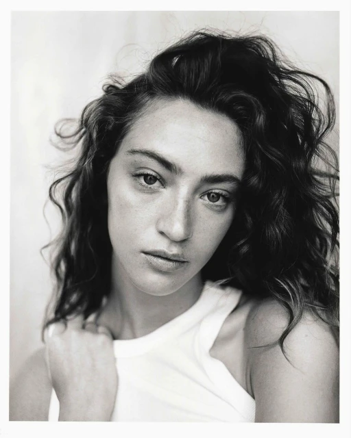 Monochrome portrait of a young adult woman with curly hair and a white top, looking at the camera against a blurred background.