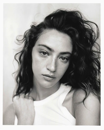 Monochrome portrait of a young adult woman with curly hair and a white top, looking at the camera against a blurred background.