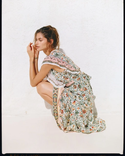 Woman with brown hair sits in front of a white wall wearing a floral print summer dress in a fashion pose.