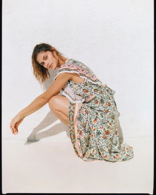 Brunette woman in floral dress crouches against a white textured wall in a studio setting. The soft light highlights the dress's pattern.