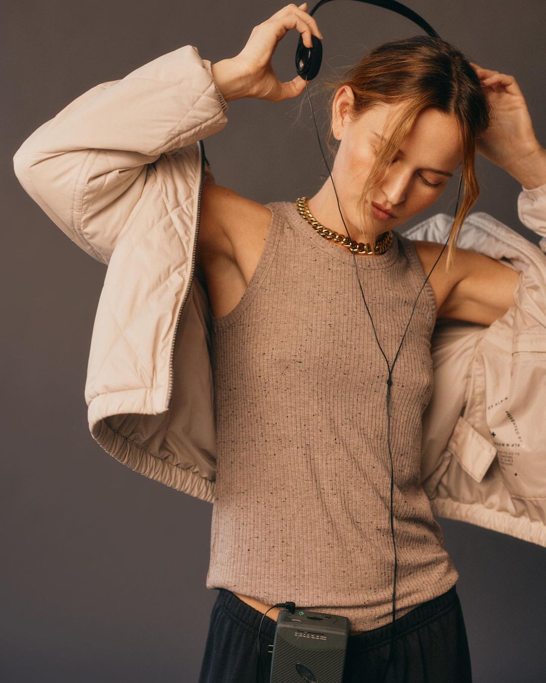 Woman in a tan top and jacket puts on headphones in a studio. She has a gold chain and an old-fashioned portable cassette player.