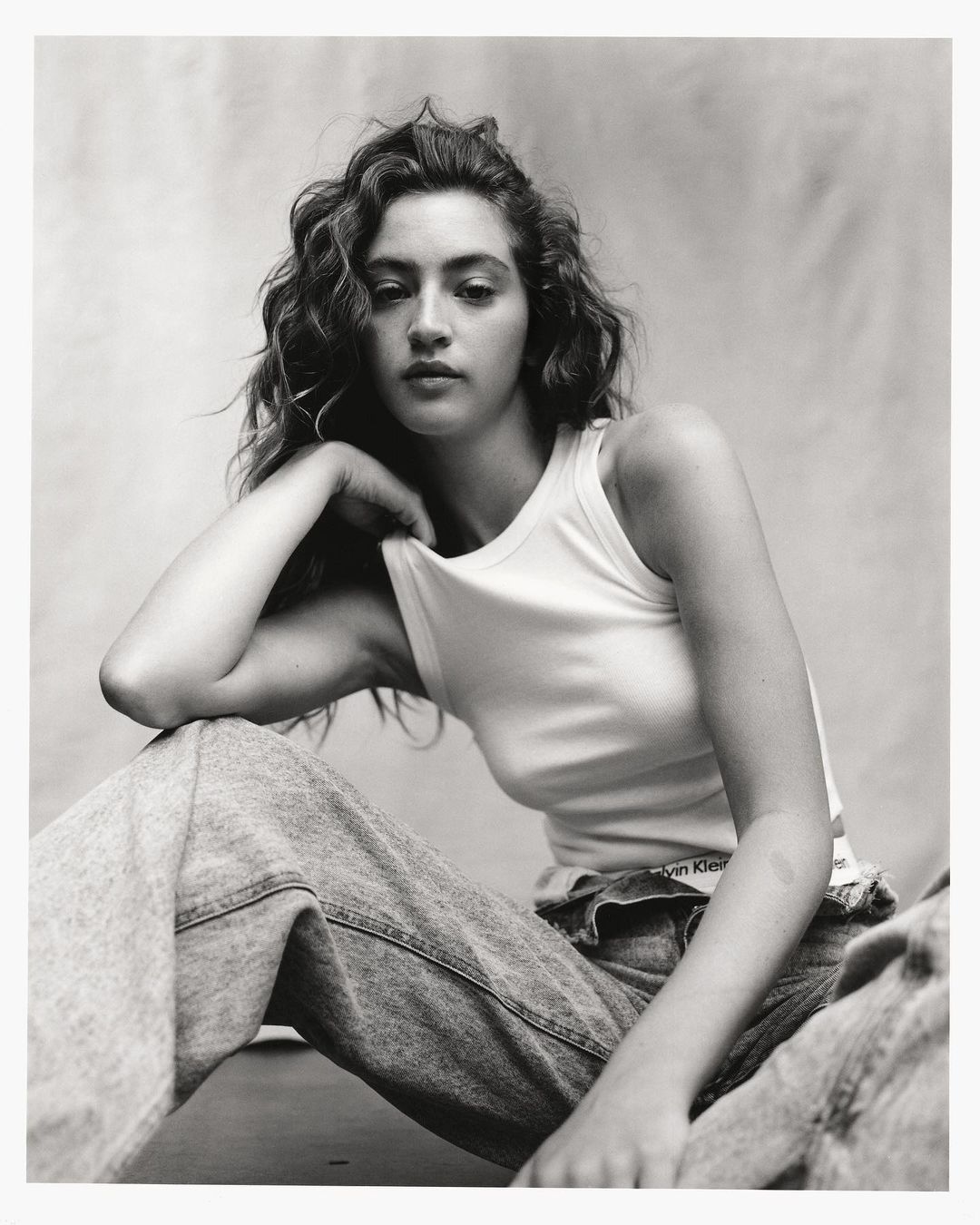 Monochrome portrait of a woman with curly hair wearing a white tank top and denim jeans against a textured backdrop.
