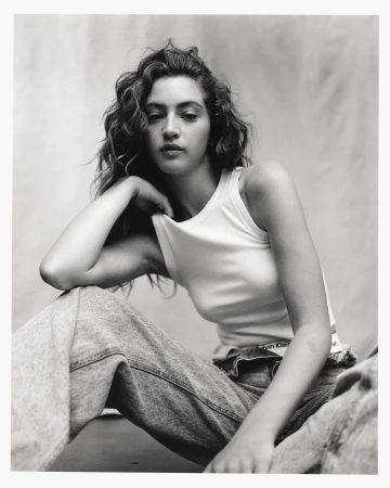 Monochrome portrait of a woman with curly hair wearing a white tank top and denim jeans against a textured backdrop.