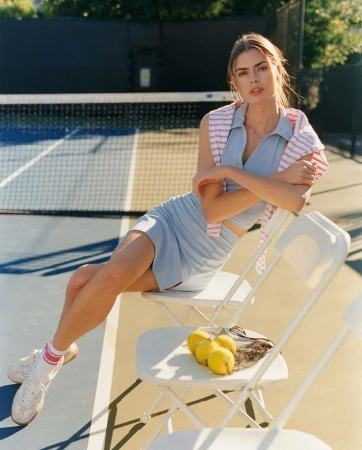 A young woman sits on a white chair on a tennis court, with lemons, wearing a blue skirt and top, with a striped sweater on her shoulders.