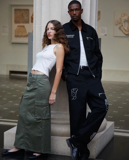 Two fashion models, one woman and one man, pose against a column inside a museum, wearing modern streetwear clothing.
