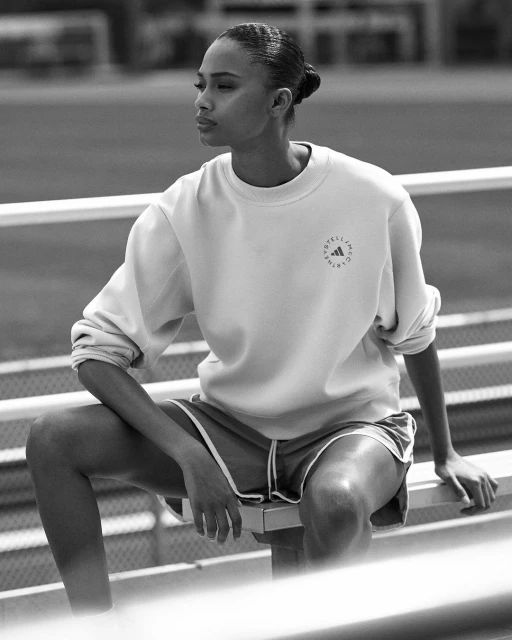 Monochrome shot of an adult woman in athletic wear sitting on bleachers on a track field, facing left, shot in daylight.