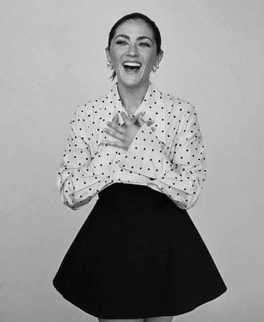 Smiling woman in polka-dot shirt and black skirt with hands on her chest.  Black and white studio portrait of a woman laughing with hands on chest.