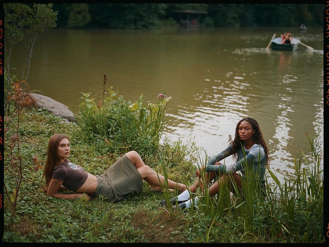 Two women pose outdoors by a lake in casual wear. One woman lies on the grass, while the other sits amongst reeds near the water's edge.