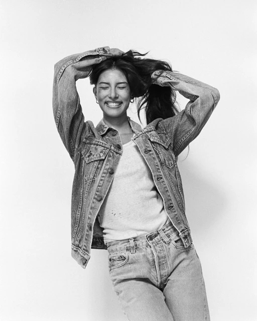 Young woman in denim jacket and jeans smiles, hands behind her head, against a white background in a studio-style fashion portrait.