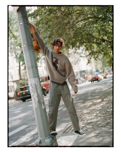 Young adult male poses next to a traffic light on a city street, wearing a sweatshirt and cargo pants on a sunny day, cars in the background.