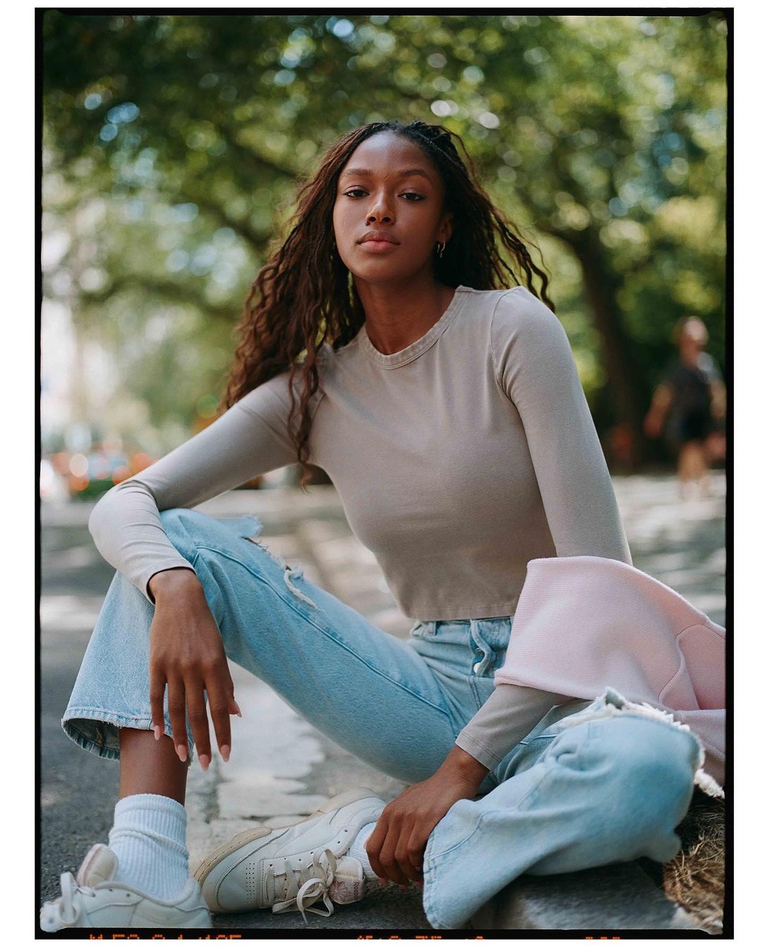 A young adult Black woman sits outdoors wearing a beige long-sleeve shirt, ripped light-wash jeans, and sneakers against a blurred background.