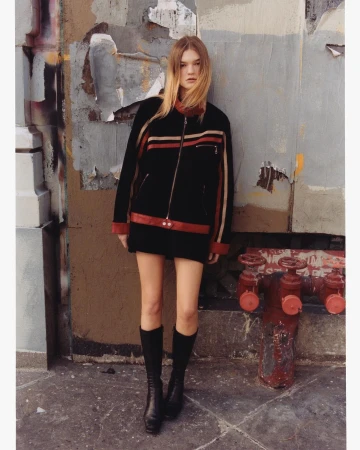 A woman in a black jacket poses in an urban setting, wearing knee-high boots with a muted color palette, standing next to a fire hydrant.