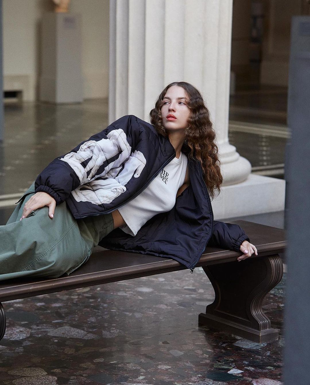 Adult female with brown curly hair poses in black jacket and green pants, reclining on a bench in a marble floored hall with columns.