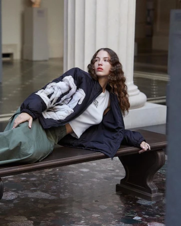 Adult female with brown curly hair poses in black jacket and green pants, reclining on a bench in a marble floored hall with columns.