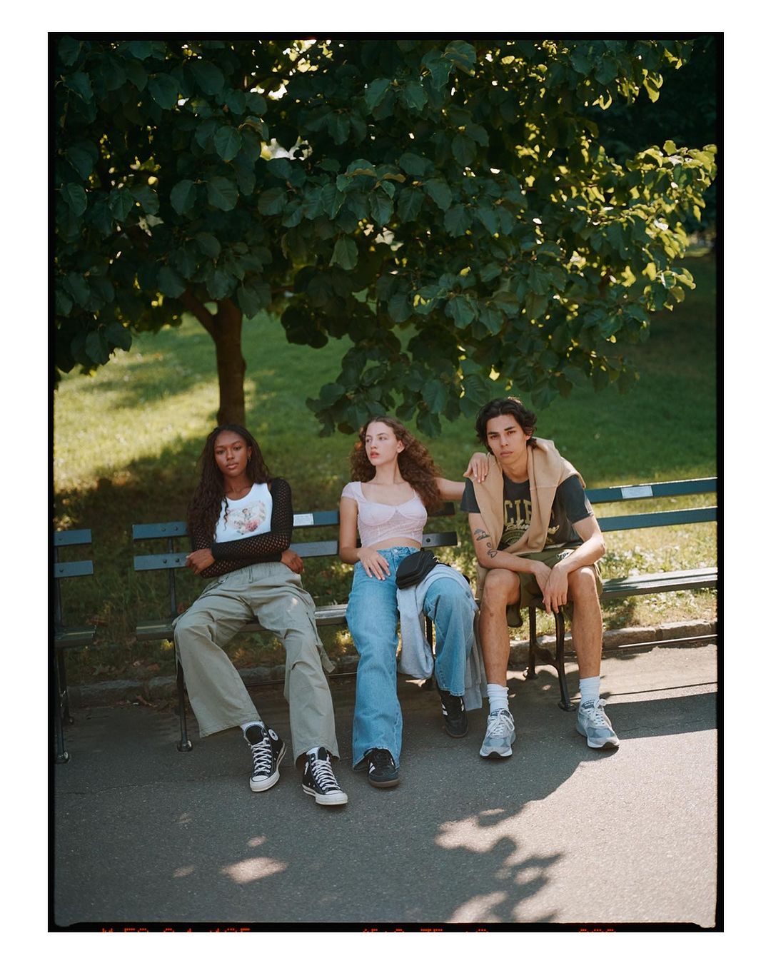 Three young adults sitting on a park bench under a tree. Two women and one man are relaxing in casual summer clothing outdoors.