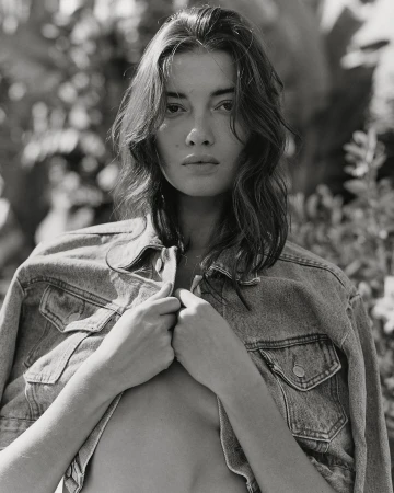 Monochrome portrait of a young adult woman in a denim jacket outdoors, with blurred foliage in the background, in soft natural light.