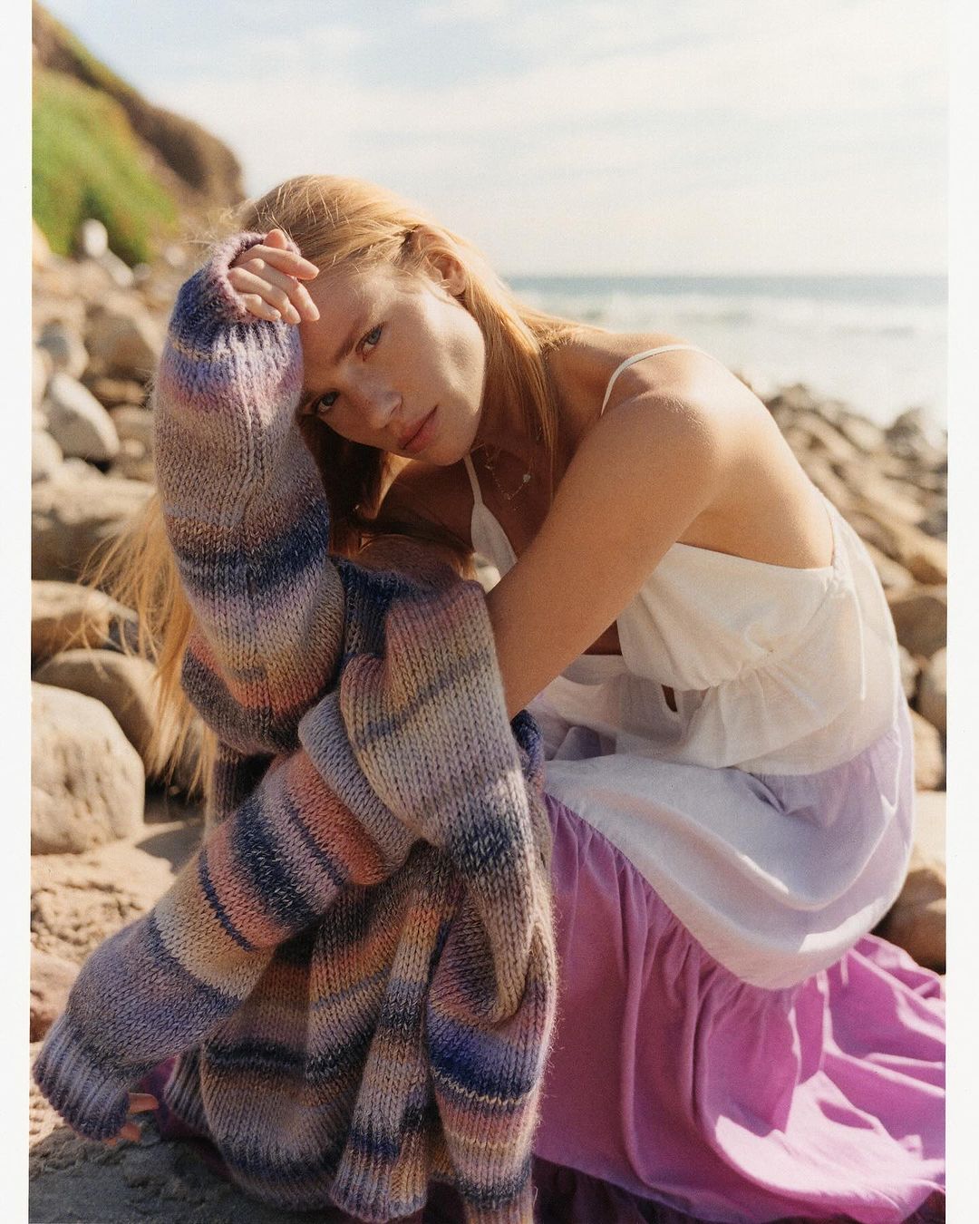 A fair-skinned woman with long red hair sits on a beach with rocks and ocean views, wearing a white dress and colorful cardigan.