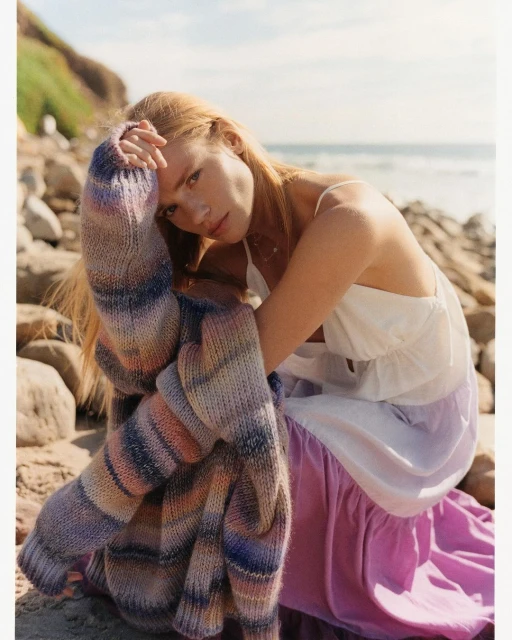 A fair-skinned woman with long red hair sits on a beach with rocks and ocean views, wearing a white dress and colorful cardigan.