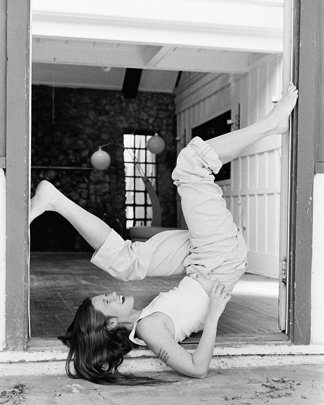 A woman smiles upside down, using a doorway for balance, wearing a white tank top and khaki pants, in a black and white photograph.