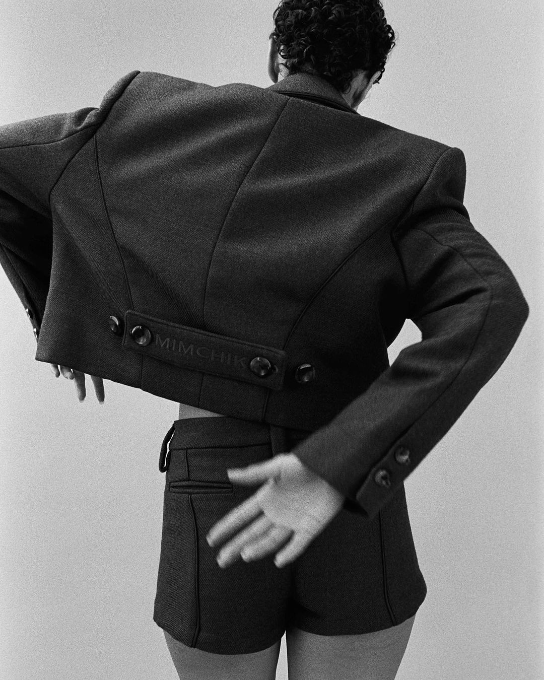 Monochrome fashion portrait from behind of a person wearing a structured blazer and shorts against a plain background