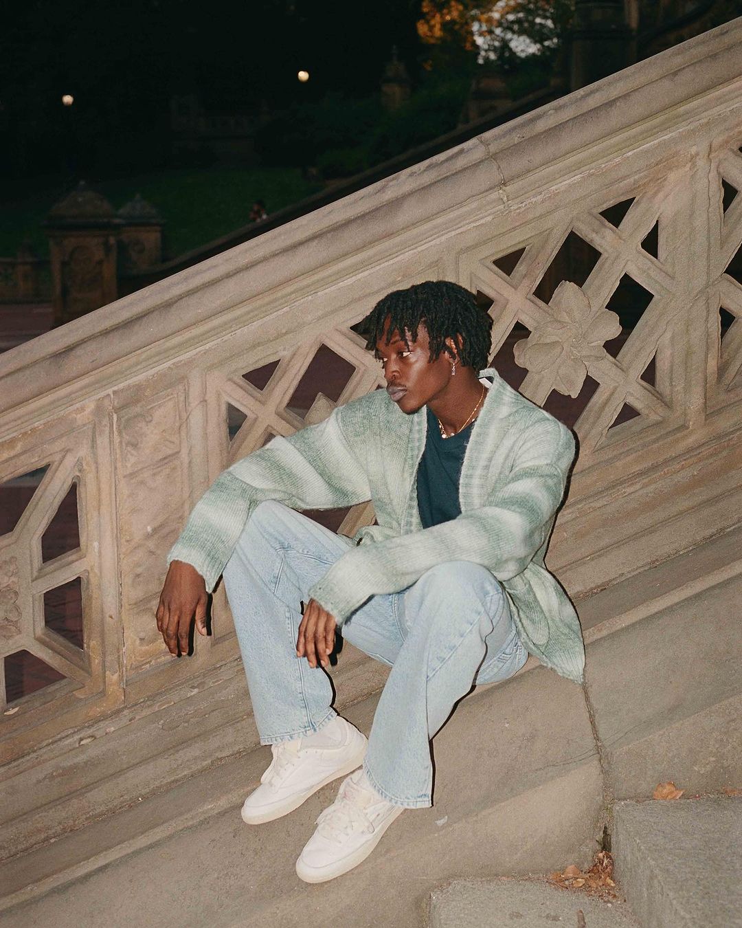 Young adult with dreadlocks sitting on stone stairs in a cardigan and jeans outdoors at night, illuminated by soft ambient light.
