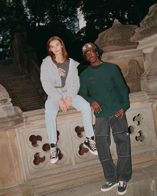 A young couple poses in stylish streetwear with a stone balustrade and trees in the background in this portrait-style outdoor shot.