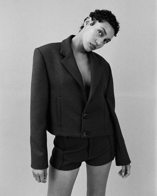 Black and white portrait of a woman with short, dark curly hair, wearing a cropped blazer and shorts, against a neutral backdrop.