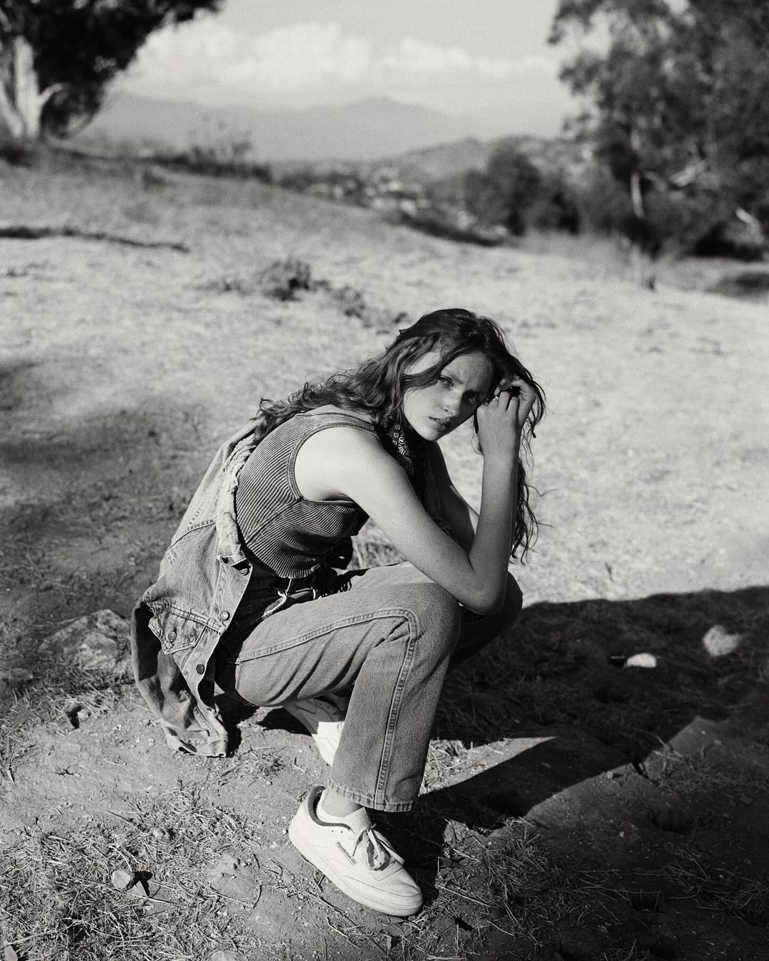 Young woman with long hair crouches in an outdoor setting, wearing a denim vest and jeans, with soft lighting and a natural background.