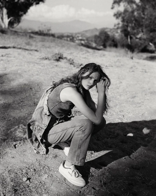 Young woman with long hair crouches in an outdoor setting, wearing a denim vest and jeans, with soft lighting and a natural background.