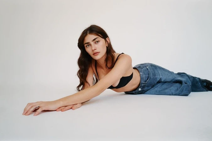 Fashion shot of a brunette adult female model in blue jeans and a black bra, reclining on a white backdrop in a photo studio.