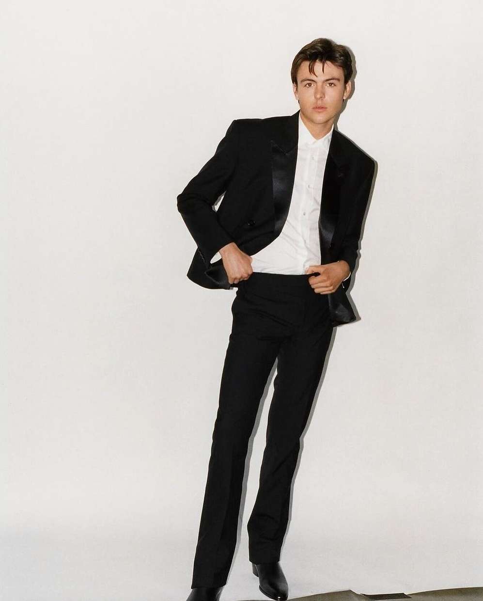 Full-length shot of a young adult man wearing a black tuxedo with a white shirt against a white background in a fashion style photograph.