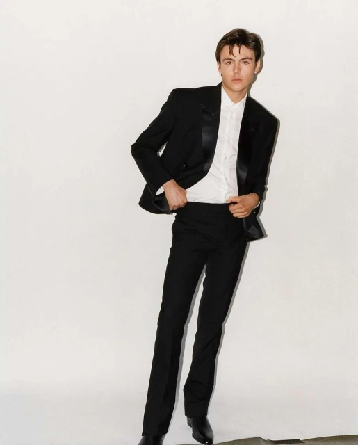 Full-length shot of a young adult man wearing a black tuxedo with a white shirt against a white background in a fashion style photograph.
