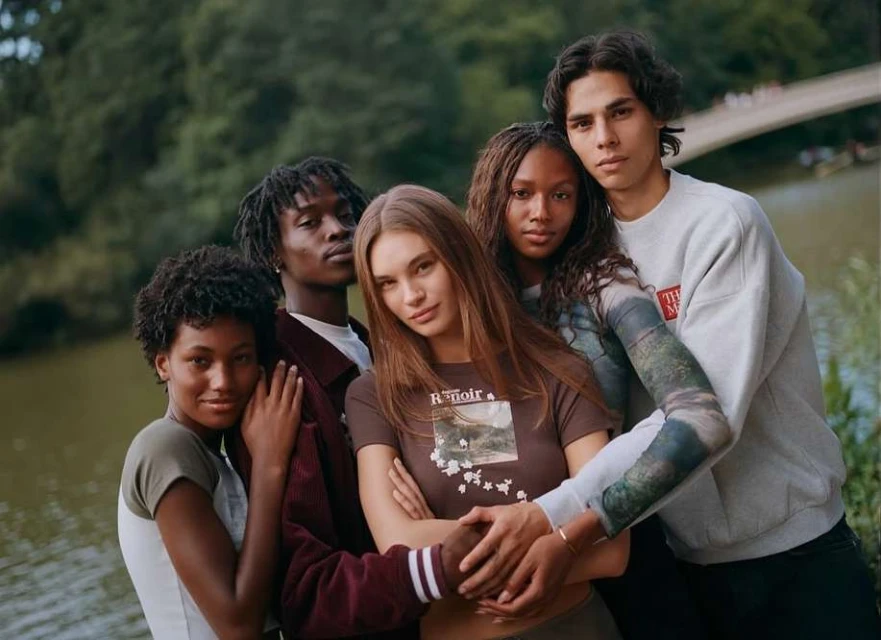 Group portrait of five diverse young adults in outdoor casual fashion; lush greenery and lake create a natural, youthful setting.