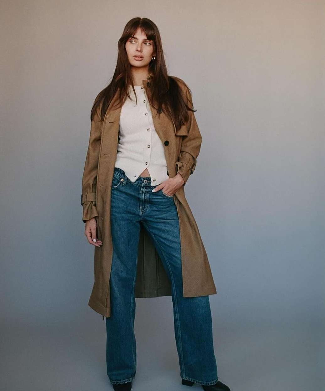 Woman with long brown hair wearing a tan trench coat, white buttoned top, and blue denim jeans against a grey backdrop