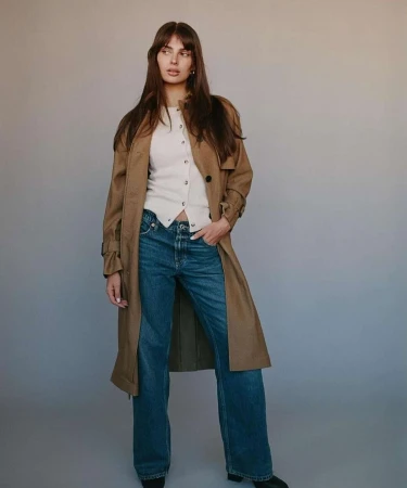 Woman with long brown hair wearing a tan trench coat, white buttoned top, and blue denim jeans against a grey backdrop