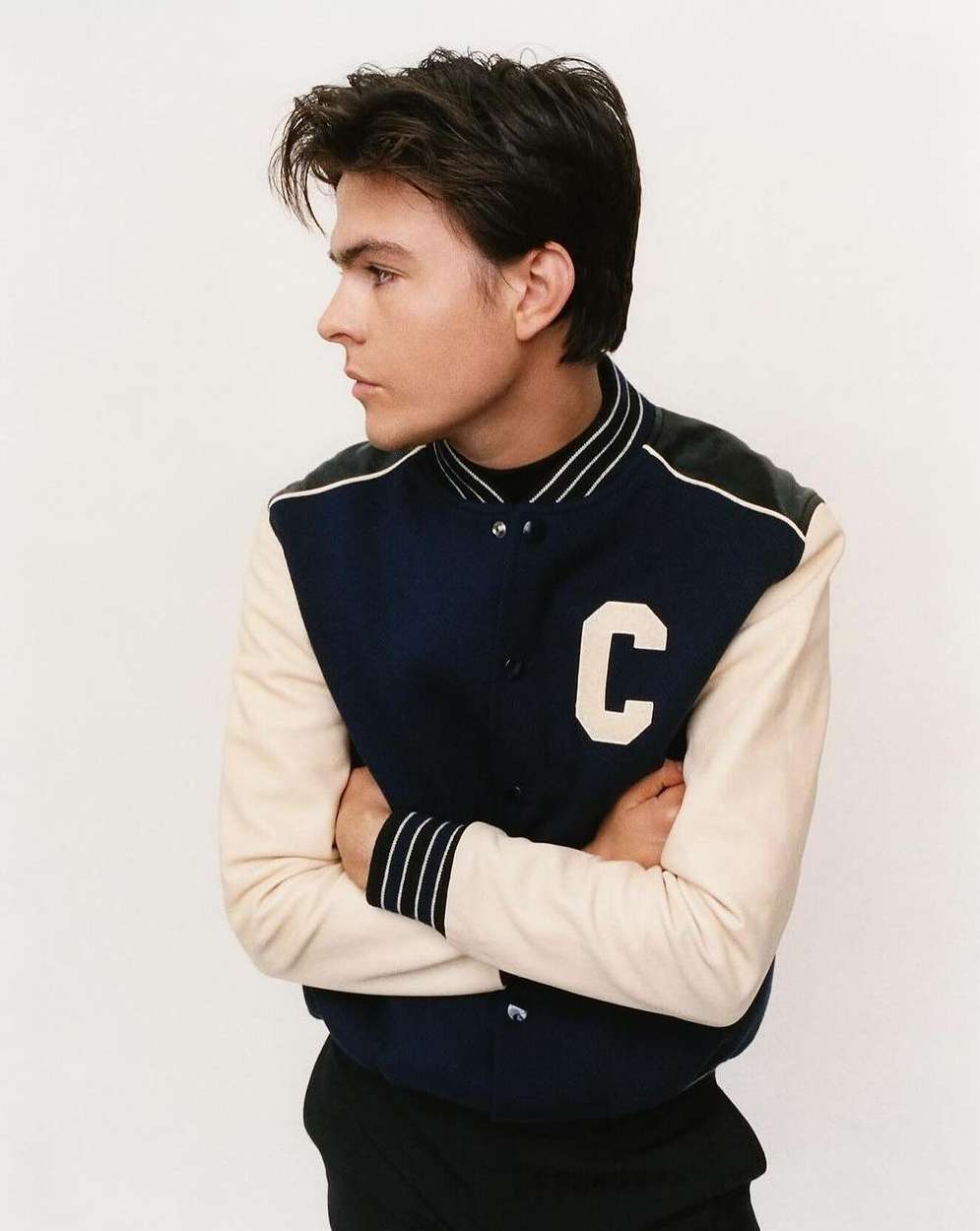 Portrait of a young adult male wearing a navy and cream varsity jacket, arms crossed, and looking off-camera against a white backdrop.